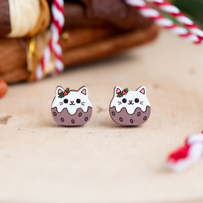 Pair of hand-painted wooden Christmas pudding cat stud earrings featuring white cats, purple-brown puddings, white icing and festive holly decorations.